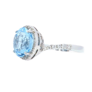 A round cut light blue gemstone ring with a silver-toned halo setting and a pavé diamond band, shown on a plain white background.